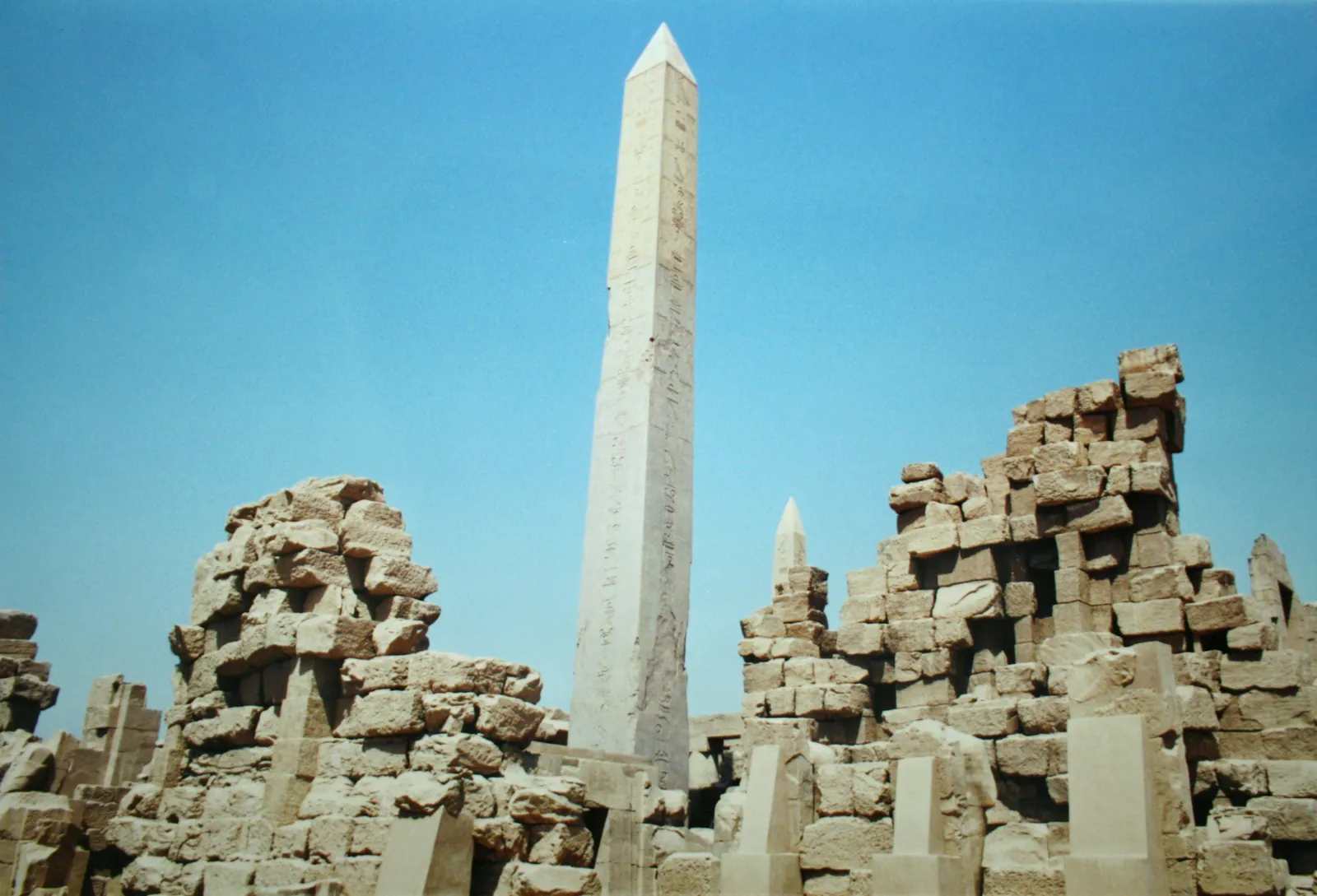 A tall granite obelisk standing among the columns and walls of the Karnak temple complex, raised by the pharaoh Hatshepsut.