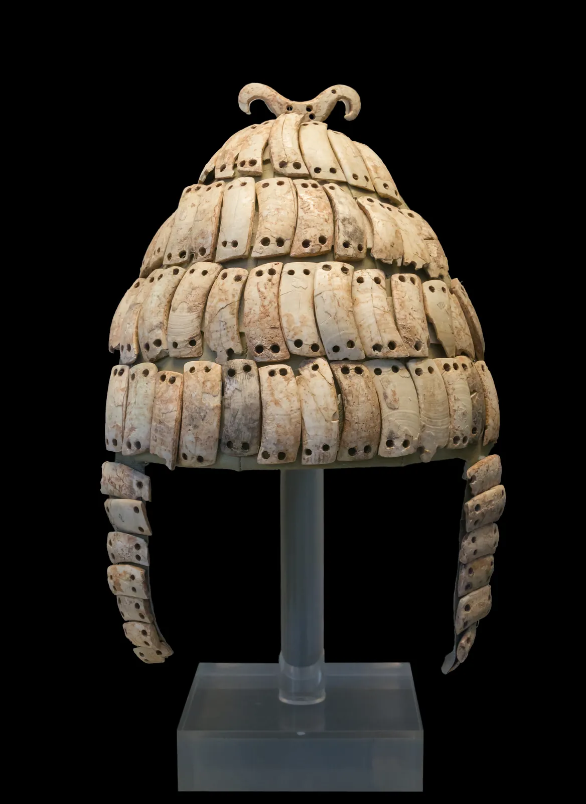 Conical helmet built from rows of curved split boar's tusks fastened to a cap, displayed in a museum case.