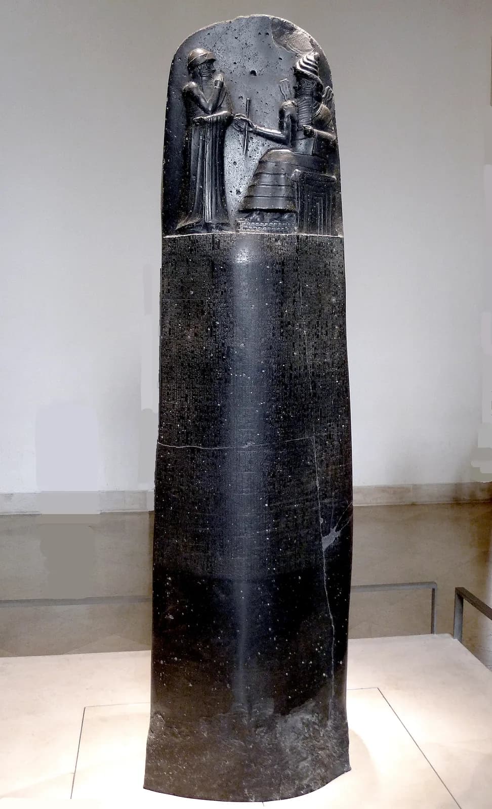 The carved upper register of the basalt stele of Hammurabi: the king standing in worship before the enthroned sun-god Shamash, who hands him the rod and ring of just rule, above the dense columns of cuneiform law.
