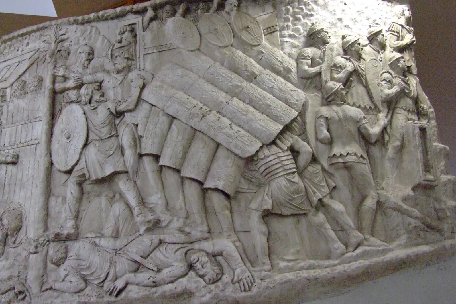 Carved relief of soldiers locking shields overhead and to the sides to form a continuous roof as they advance.