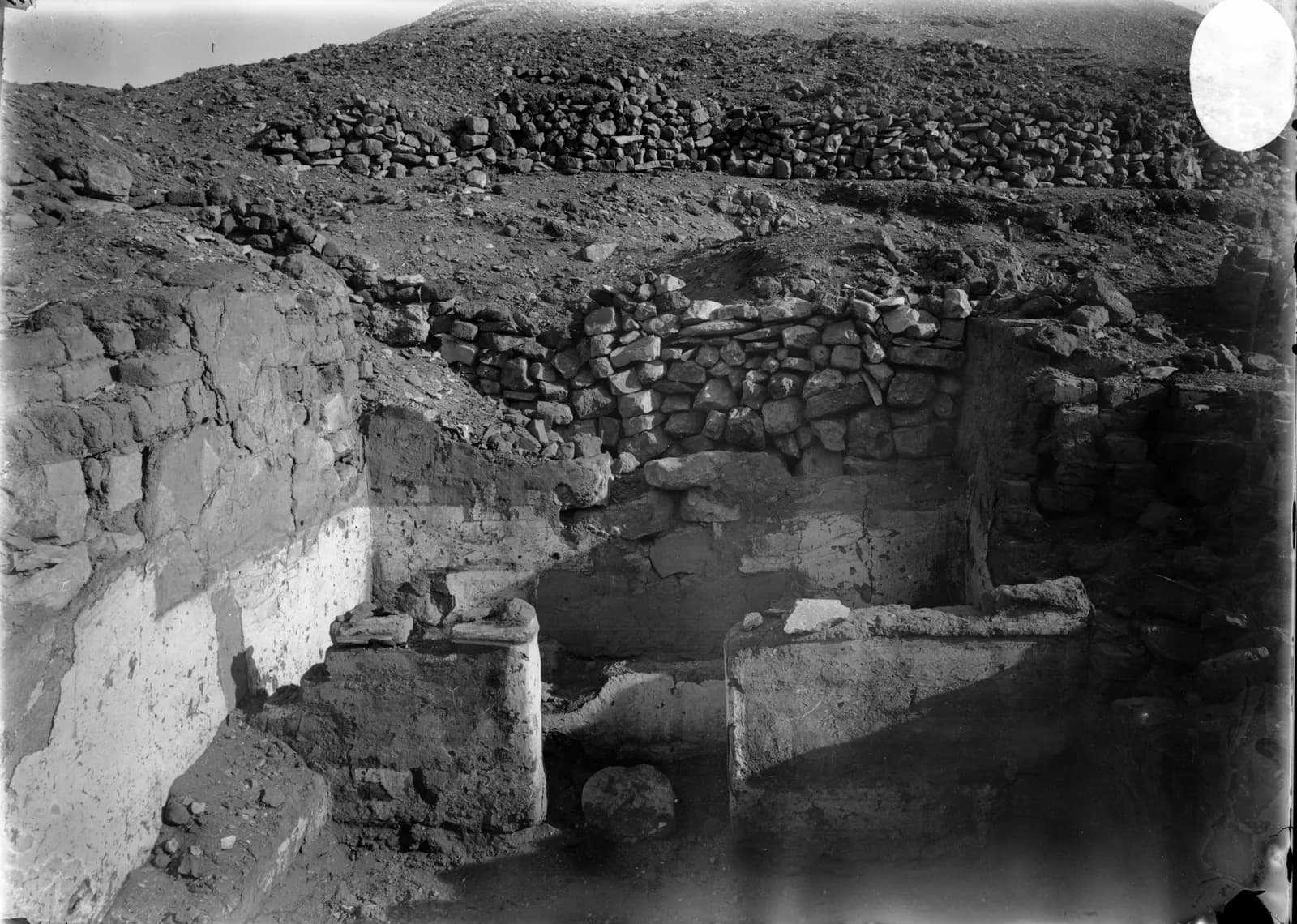 A historical photograph of an excavation in the desert: workers among low mudbrick walls and spoil heaps below a rocky hillside.