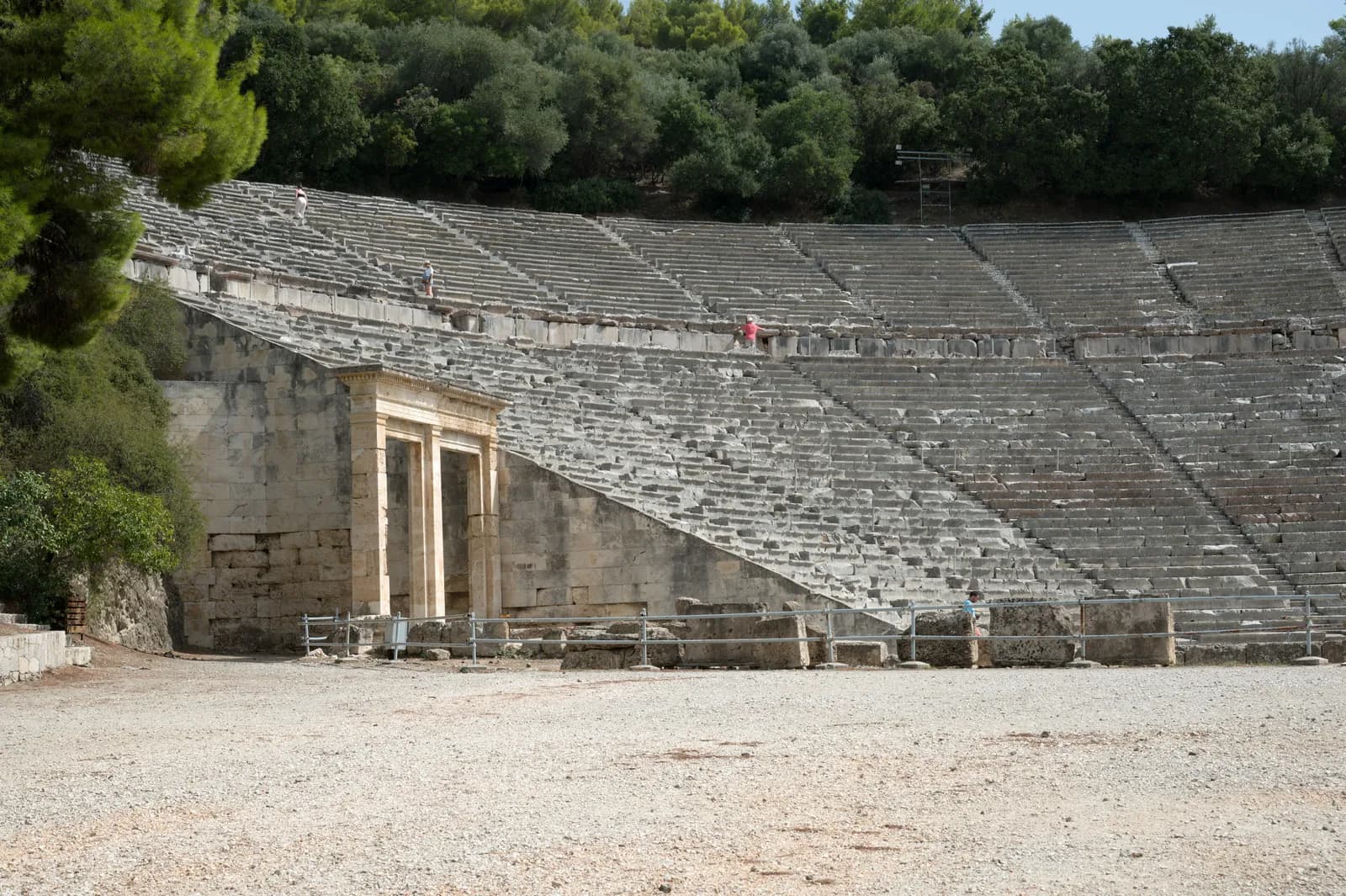 A large Greek theatre of tiered stone seating in a wide arc around a circular orchestra, set into a hillside with wooded hills behind.
